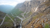 A paved road zig zags down a hillside, over a waterfall and into a valley with steep walls